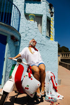 A woman wearing a white t-shirt, blue shorts, and a red-and-white patterned headscarf sits on a white, red, and green scooter decorated with cherry-patterned covers, set against a light blue Mediterranean-style building under a clear blue sky.