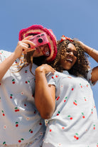 Two women outdoors under a clear blue sky, wearing matching cherry print pjs. One holds a pink camera to her eye.