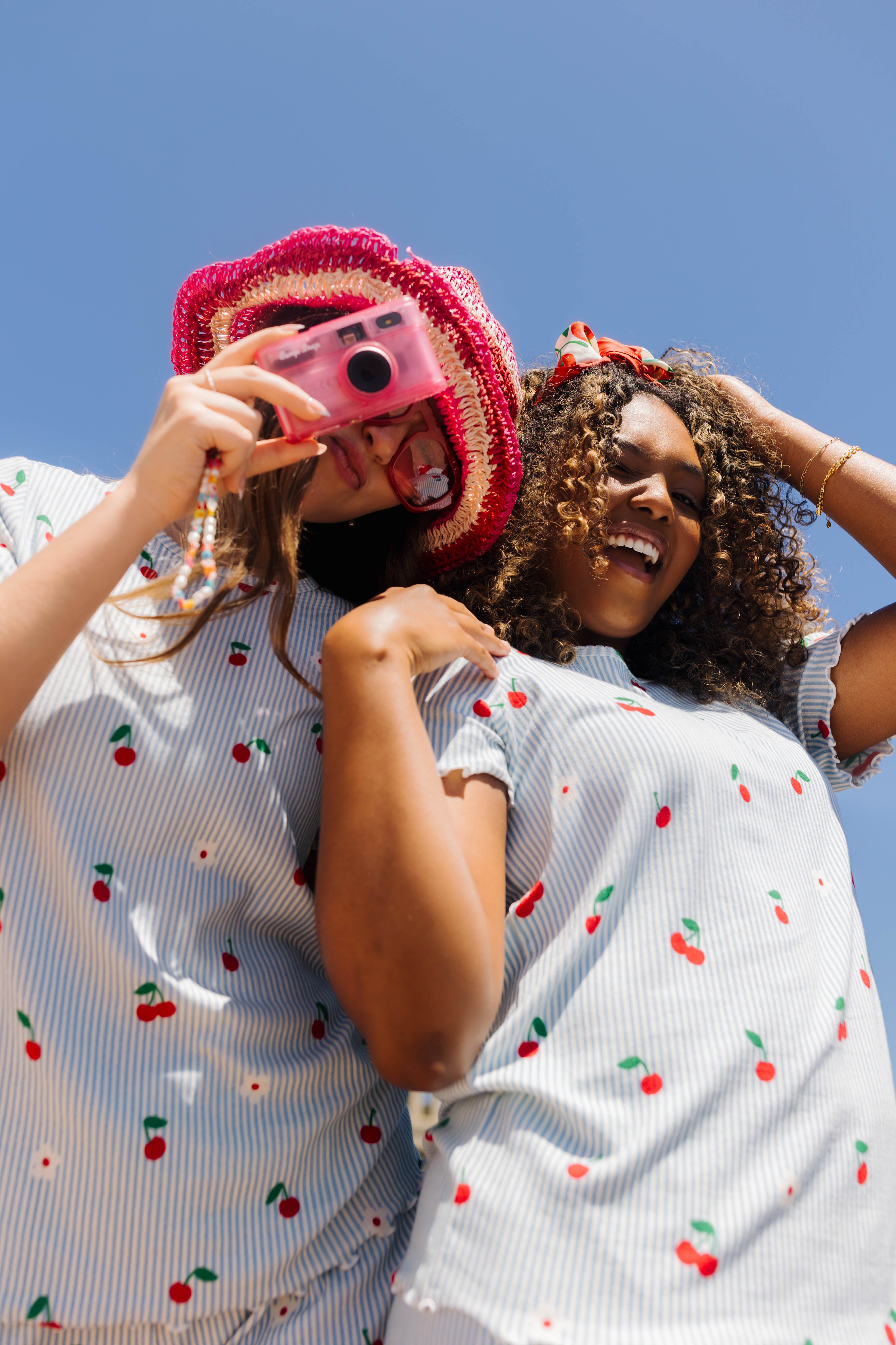 Two women outdoors under a clear blue sky, wearing matching cherry print pjs. One holds a pink camera to her eye.