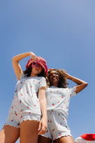 Two women wearing matching white short-sleeve pajama sets with a red and green cherry print, posing against a clear blue sky.