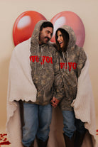 Two people wearing matching camouflage hoodies with 'love you' text, standing against a beige wall with heart-shaped balloons and wrapped in the scalloped blanket.