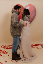 Couple embracing in front of heart-shaped balloons with red rose petals on a beige background with Krista wrapped in the blanket