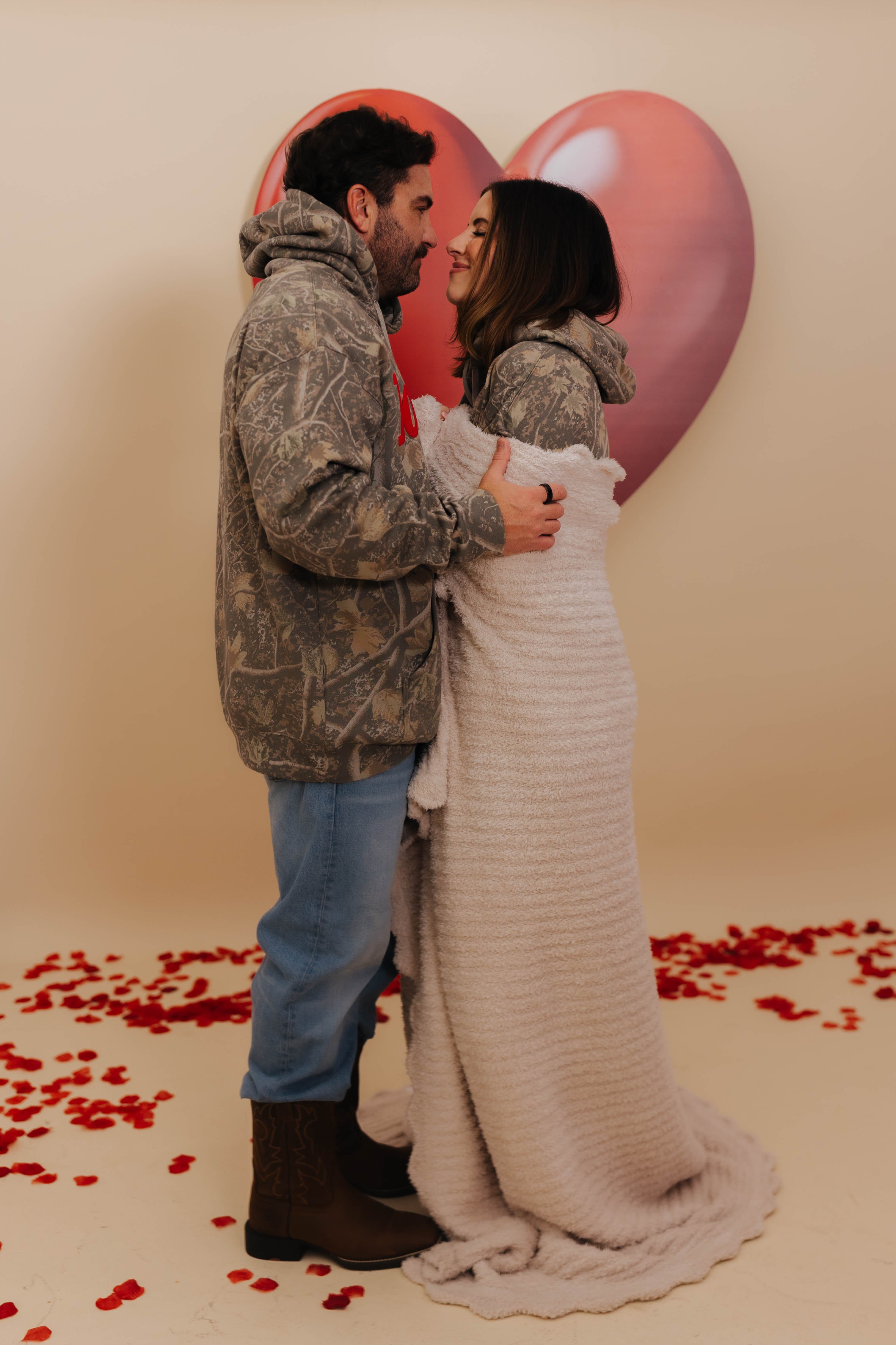 Couple embracing in front of heart-shaped balloons with red rose petals on a beige background with Krista wrapped in the blanket