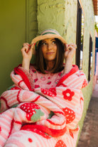 A woman wrapped in a pink and red strawberry-patterned plush blanket, wearing a straw hat, leaning against a textured olive green wall.