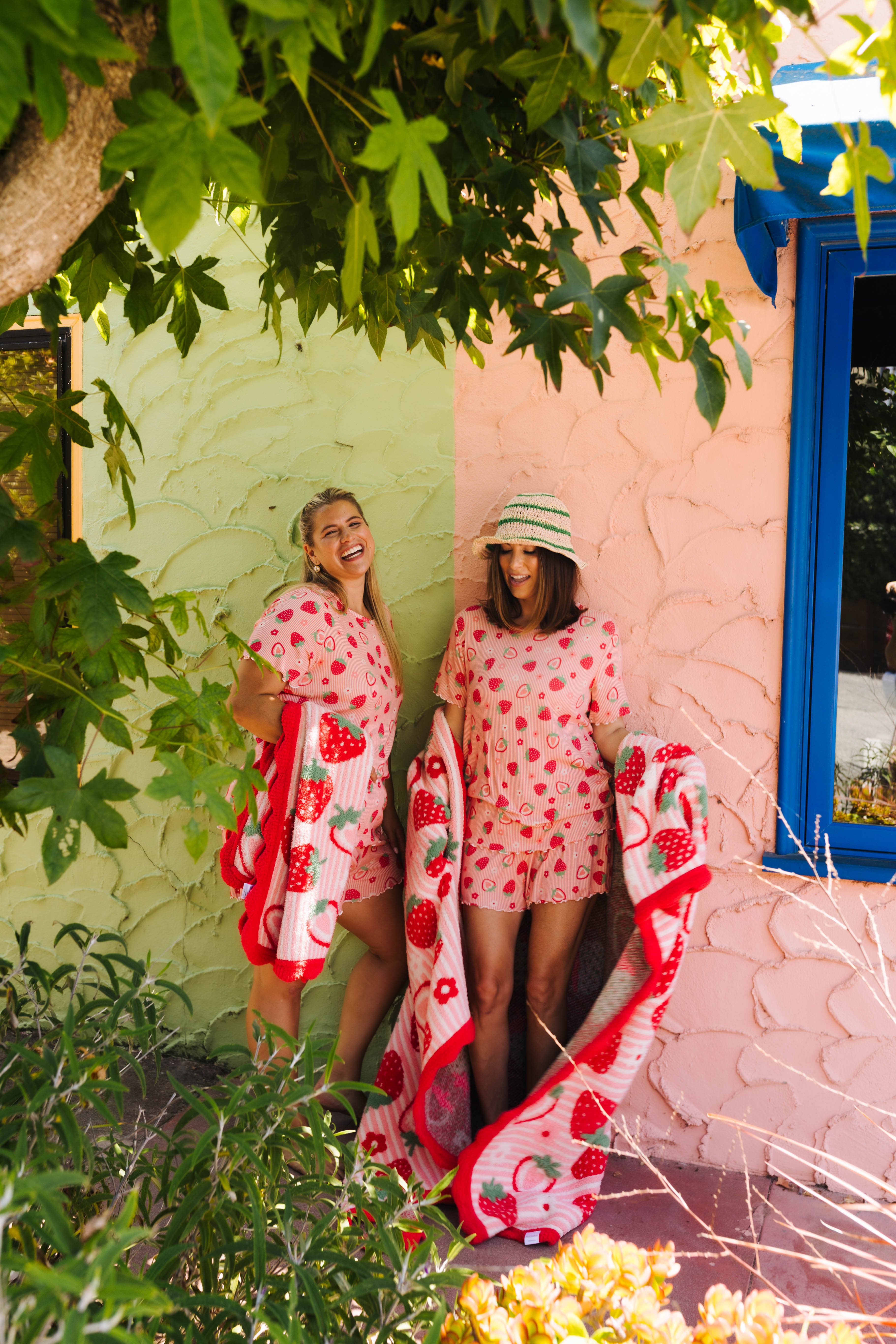 Two women standing outdoors in front of a pink and green stucco wall, wearing matching strawberry pjs and holding matching red-and-white strawberry blankets.