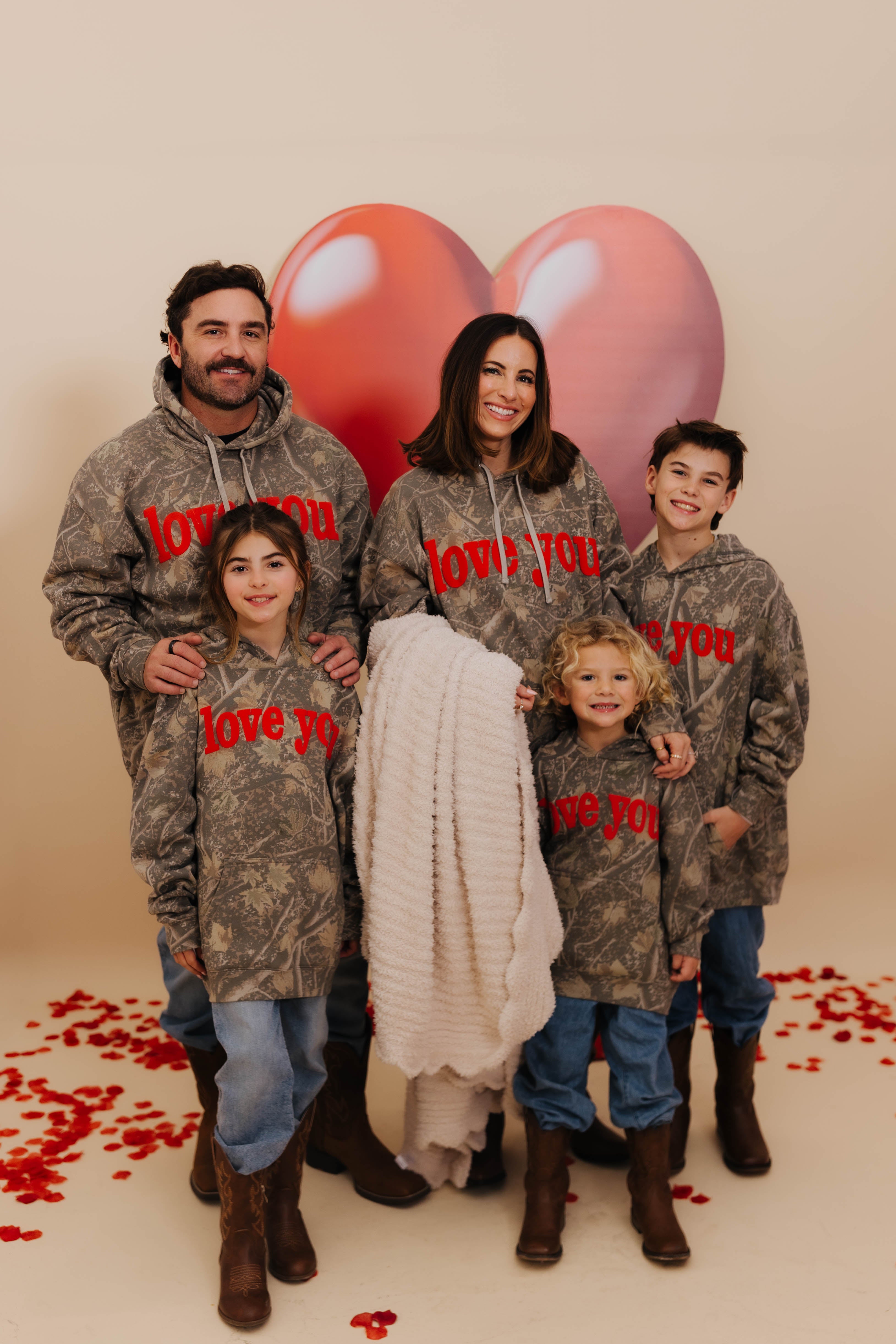 Family of five wearing matching camouflage hoodies with 'love you' text, standing in front of heart-shaped balloons. Krista holding the blanket.