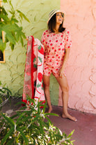 A woman wearing a pink strawberry-print short-sleeve pajama set and holding a matching strawberry-patterned blanket, posing outdoors against a split green and pink stucco wall.