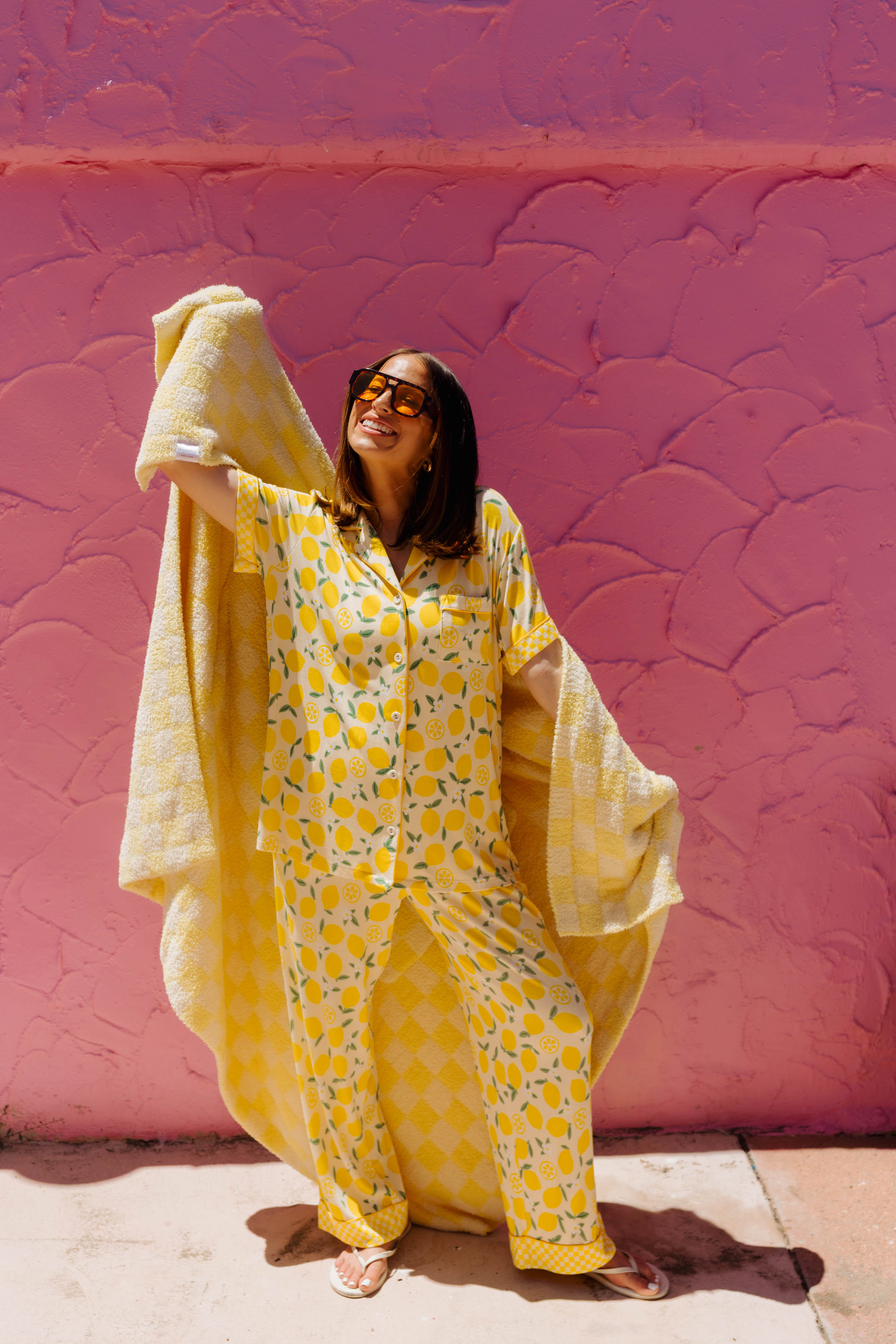 A woman wearing a yellow patterned pj set stands against a textured pink wall, holding a matching yellow blanket draped over her arm.