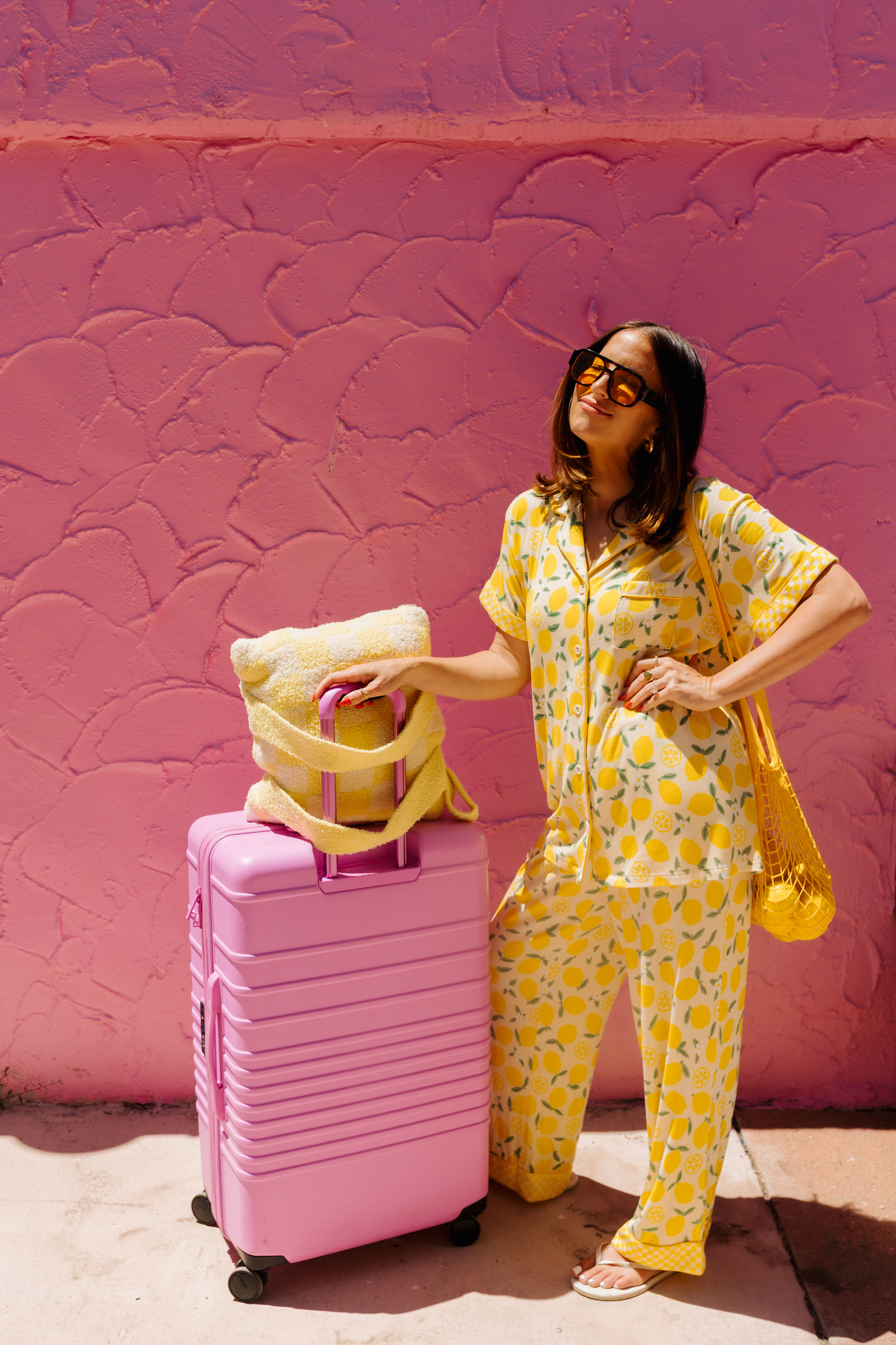 A woman wearing a yellow and white lemon-patterned pajama set stands next to a pink suitcase and a pink pillow against a textured pink wall.