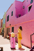 A woman in a yellow lemon pajama set stands next to a pink rolling suitcase on a sunny sidewalk beside a bright pink stucco building.