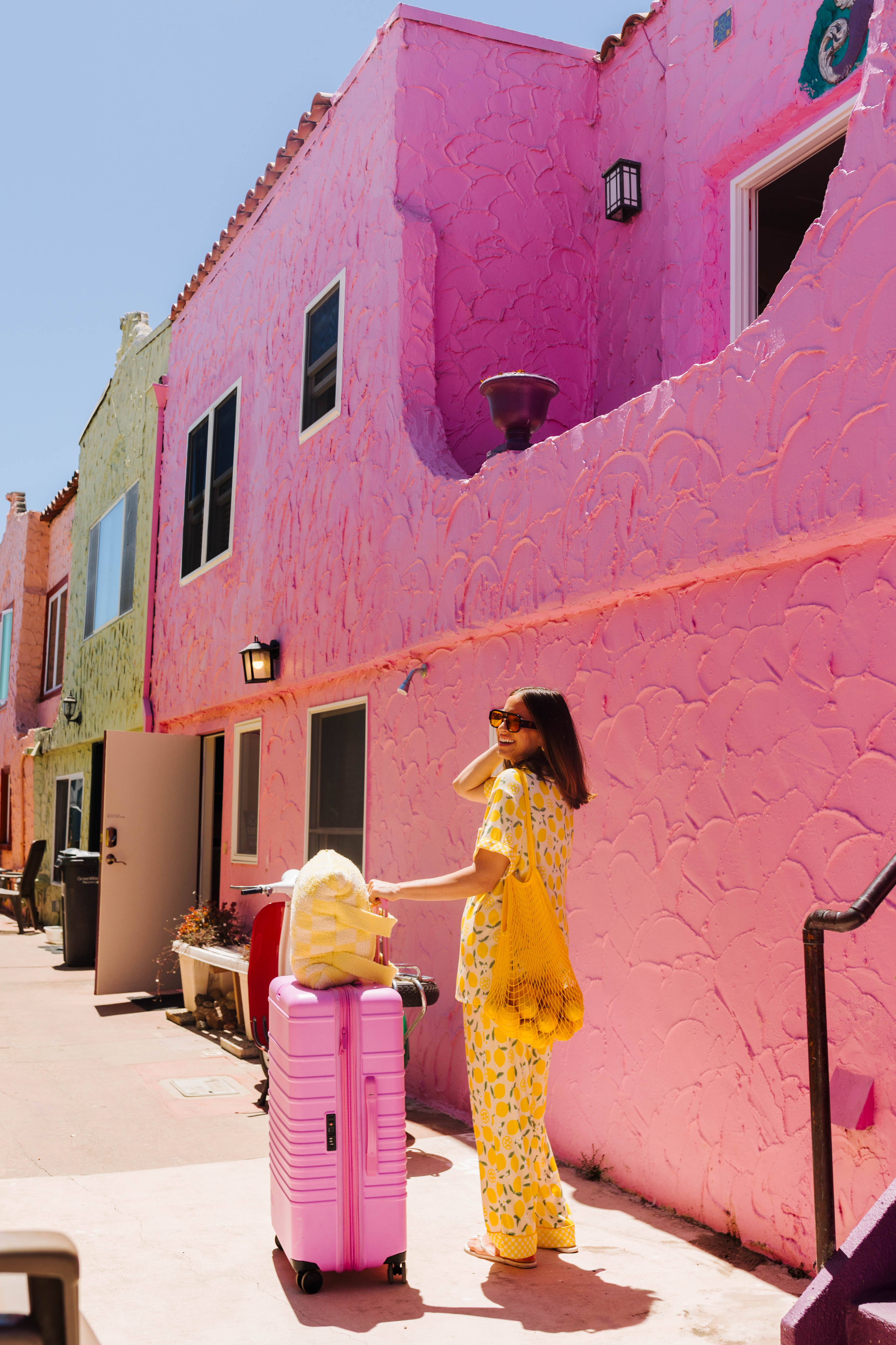A woman in a yellow lemon pajama set stands next to a pink rolling suitcase on a sunny sidewalk beside a bright pink stucco building.