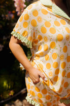 A close-up of a person wearing a short-sleeve collared top or dress with an allover orange and green citrus print, photographed outdoors with blurred foliage in the background.