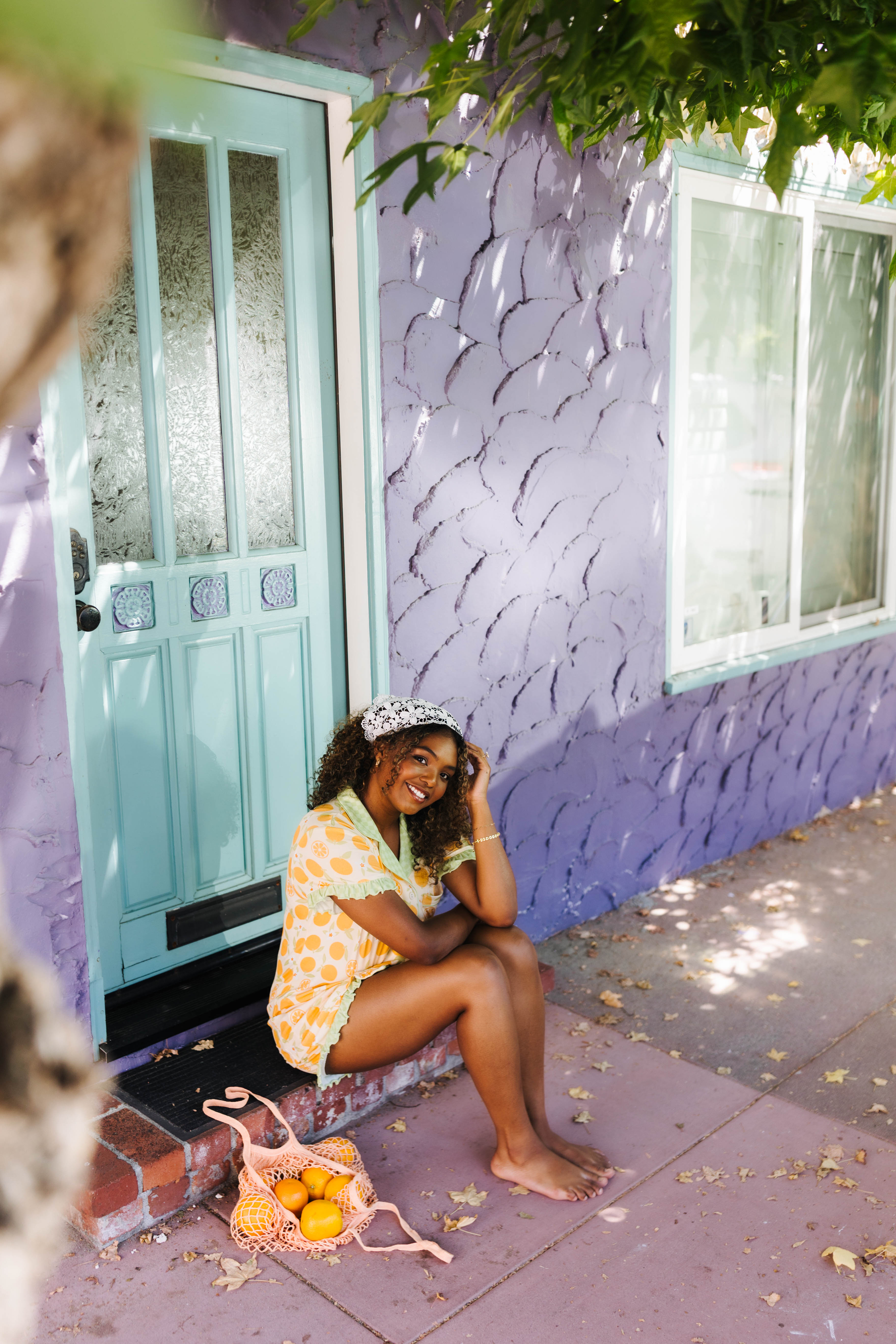 A woman sits barefoot on the steps of a purple house with a turquoise door, wearing an orange pj set. A pink reusable bag with oranges sits on the sidewalk beside her.