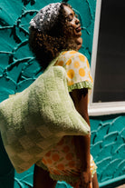 A woman wearing orange pjs carrying a large textured  green quillow posed against a vibrant teal textured wall.