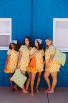 Four women posing together in matching yellow and orange patterned pjs standing barefoot against a bright blue stucco wall with windows.