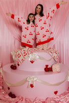 Two people in cherry-patterned wearable standing on a pink cake with heart decorations.