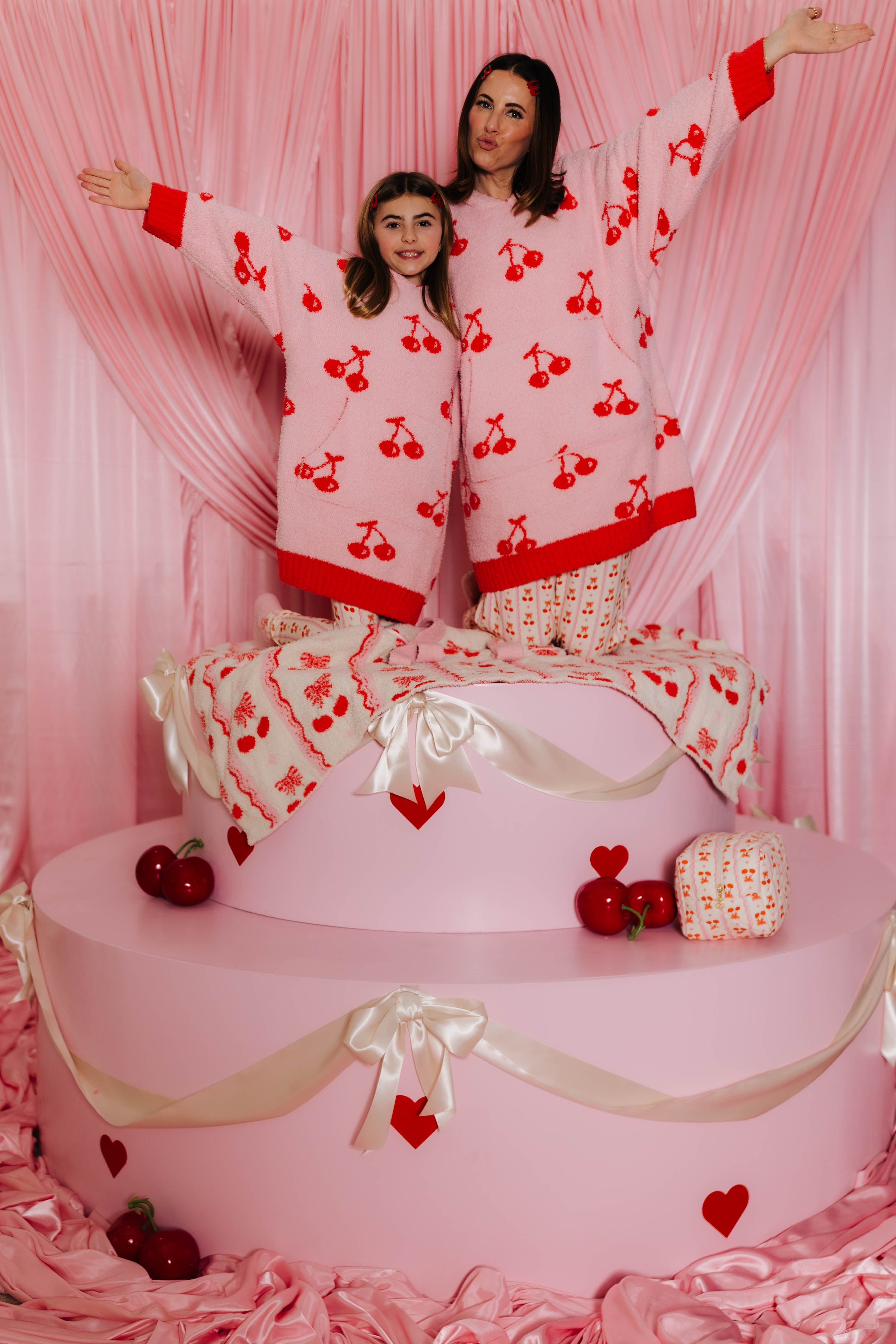 Two people in cherry-patterned wearable standing on a pink cake with heart decorations.