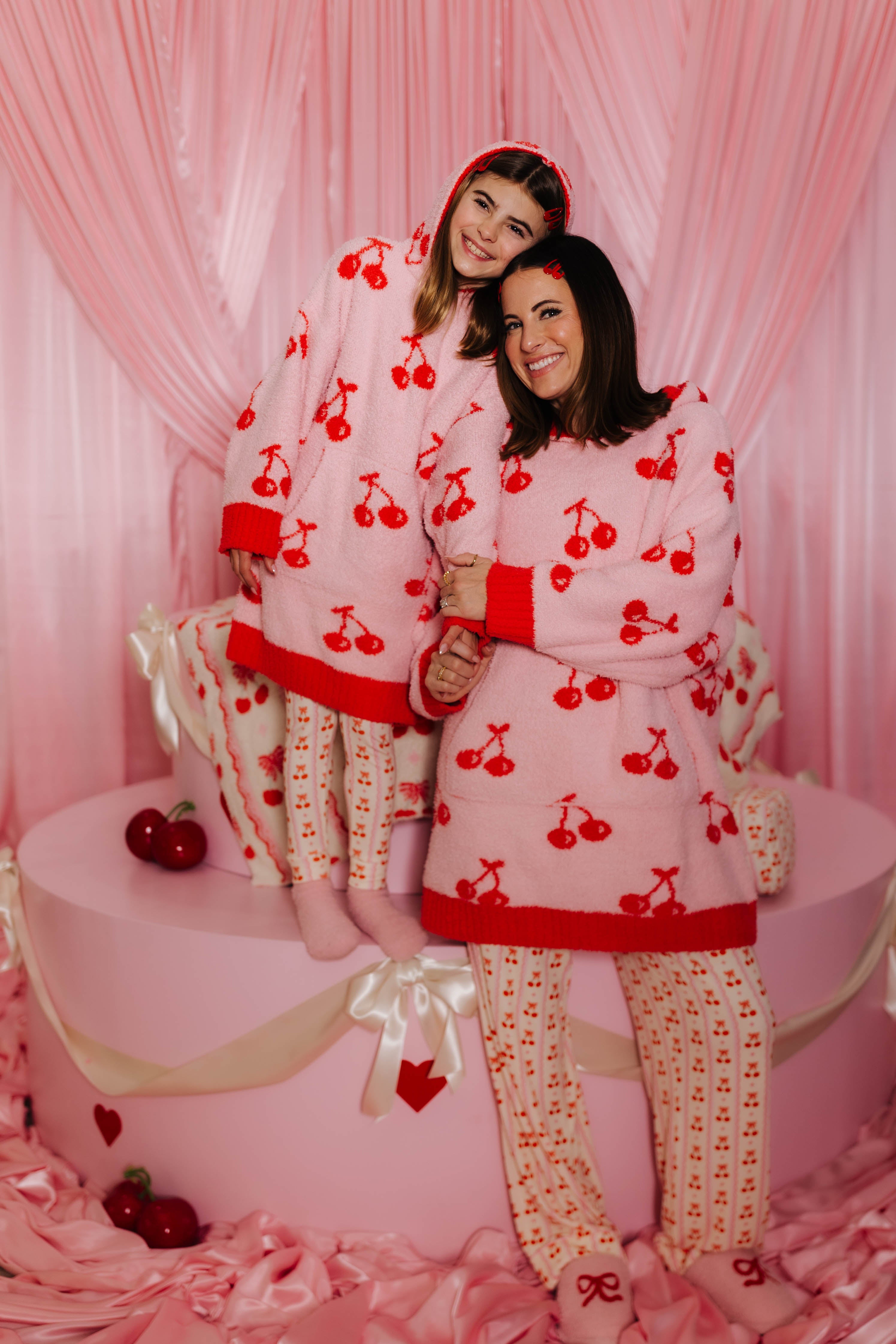 Krista and Kolly in matching cherry-patterned wearables standing in front of a pink curtain.