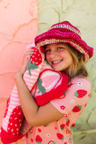 A smiling young girl wearing a pink and red strawberry-themed knit hat and matching strawberry-print top, hugging a pink and red strawberry knit blanket against a split peach and green textured wall.