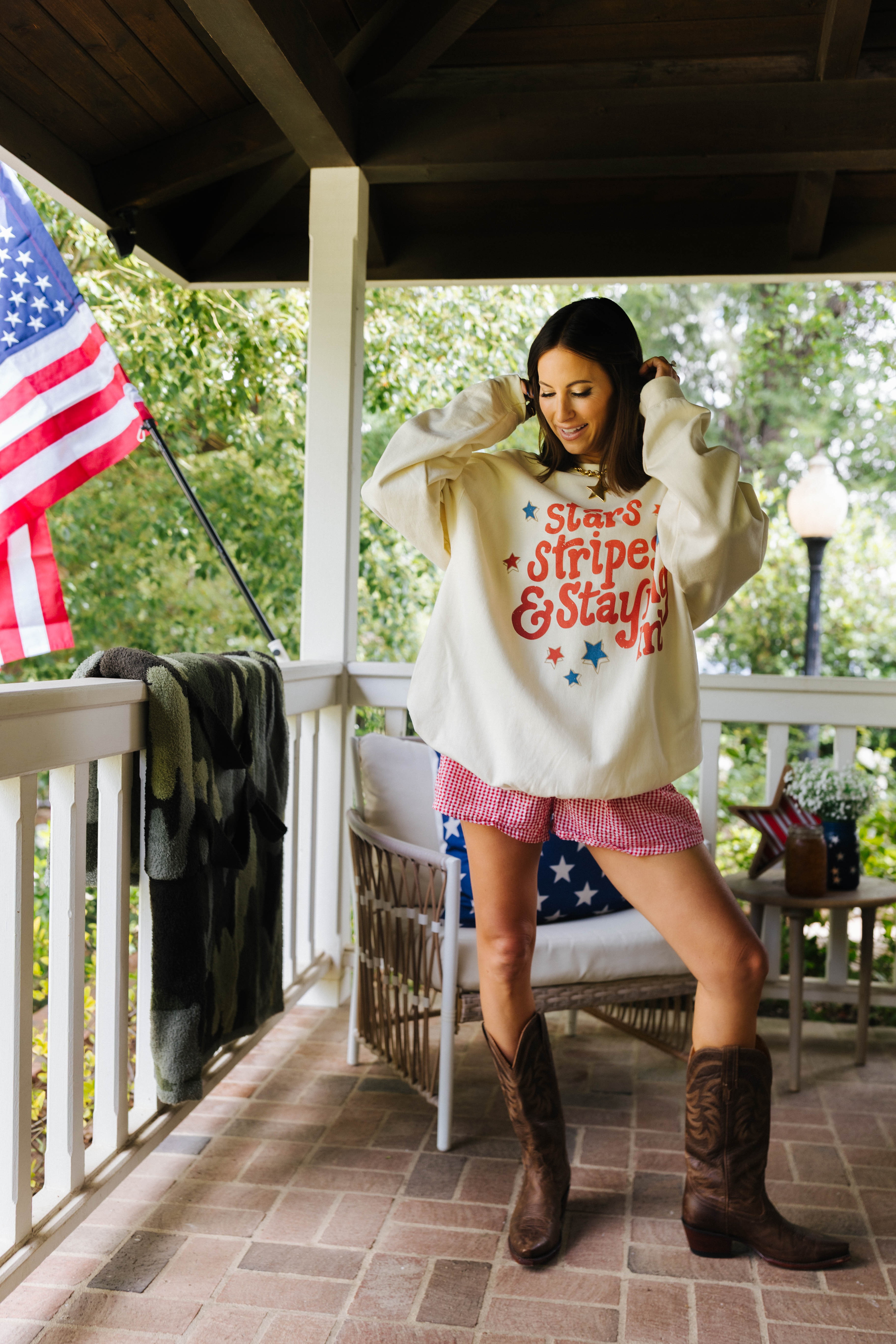 Woman on a porch wearing a sweatshirt with text and pink shorts, standing next to an American flag.