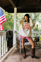 Woman on a porch wearing a sweatshirt with text and pink shorts, standing next to an American flag.