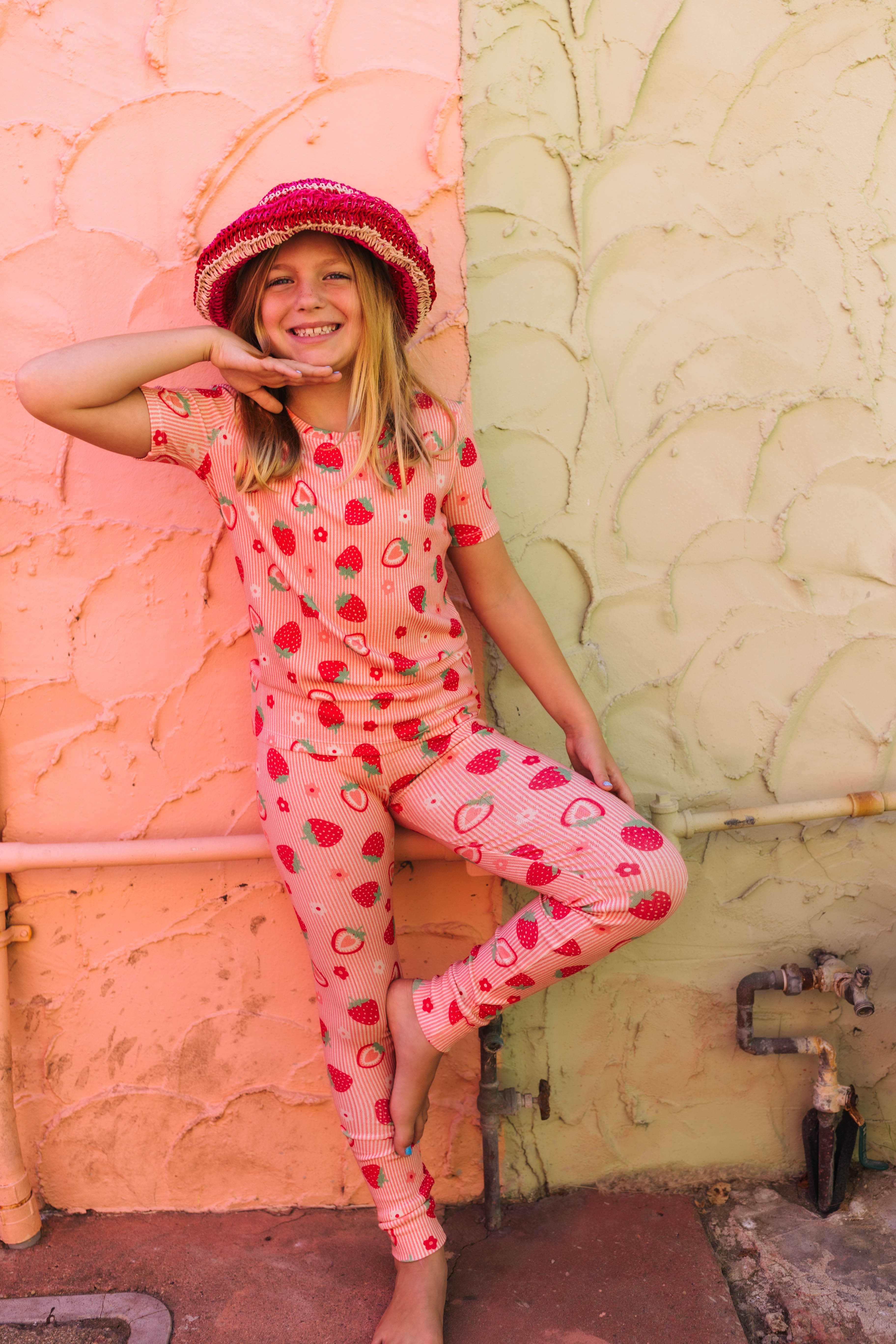 A young girl smiling and posing in a strawberry-print pajama set with a matching red hat, leaning against a split peach and beige stucco wall.