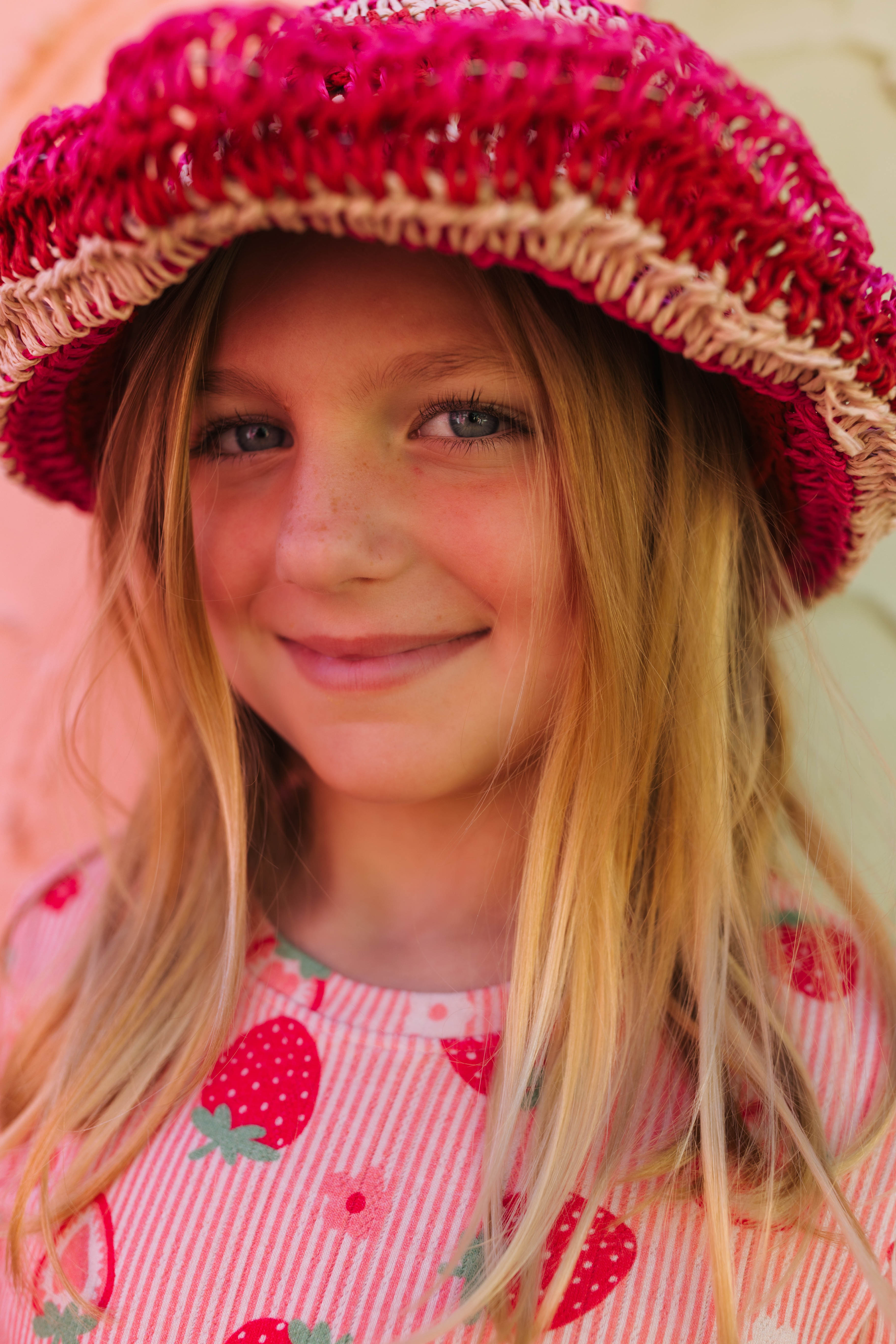 A young girl with long blonde hair smiling while wearing a bright pink and red striped knit hat with a straw-like trim and a pink strawberry-patterned top.