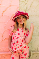 A young girl smiling while wearing a pink strawberry-patterned short-sleeve top and matching pants, along with a pink woven hat, standing against a split pink and beige textured wall.