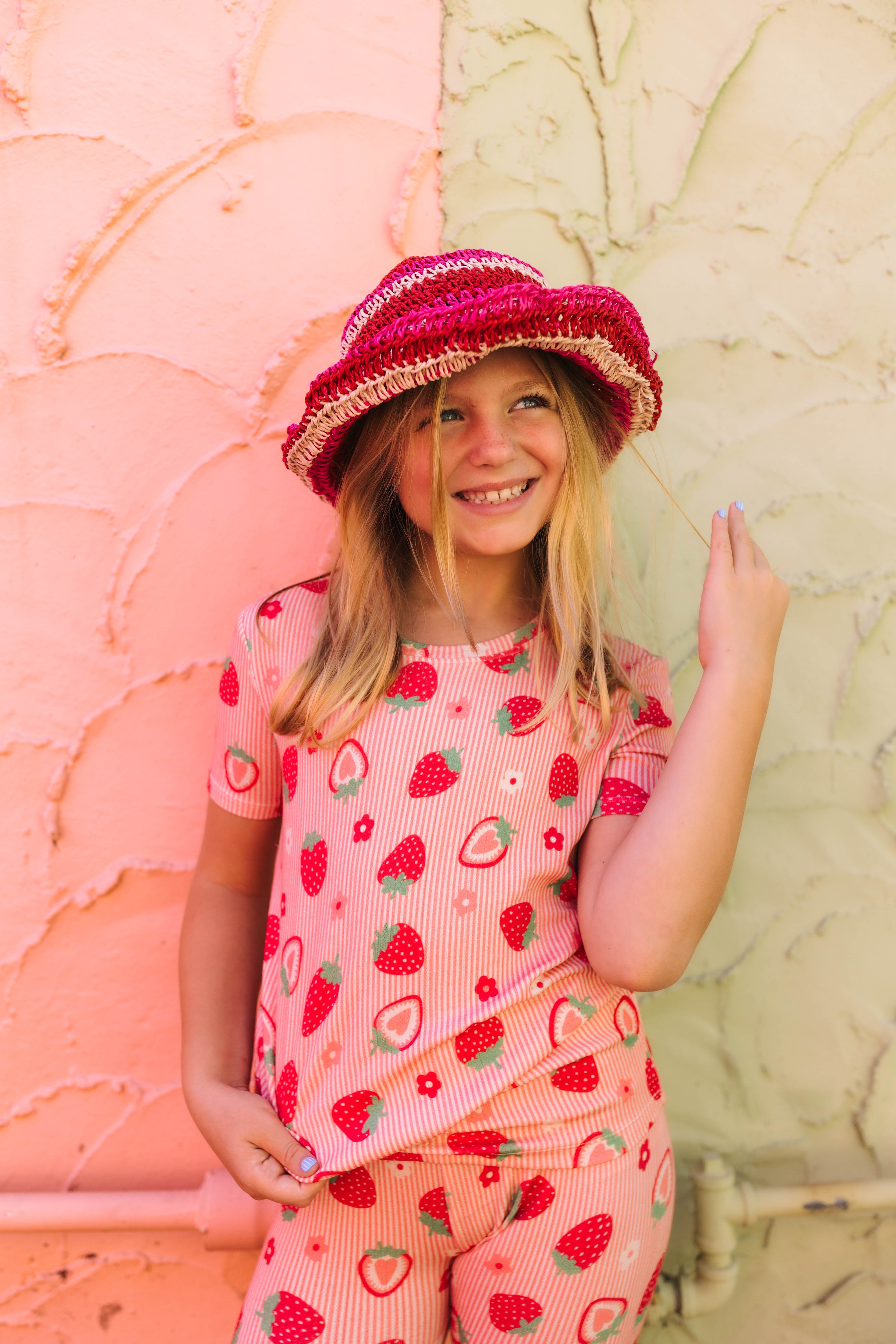 A young girl smiling while wearing a pink strawberry-patterned short-sleeve top and matching pants, along with a pink woven hat, standing against a split pink and beige textured wall.