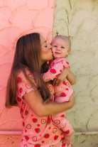 A woman and a baby wearing matching pink strawberry-patterned pajamas, with the woman kissing the baby on the cheek against a split pink and beige textured wall.