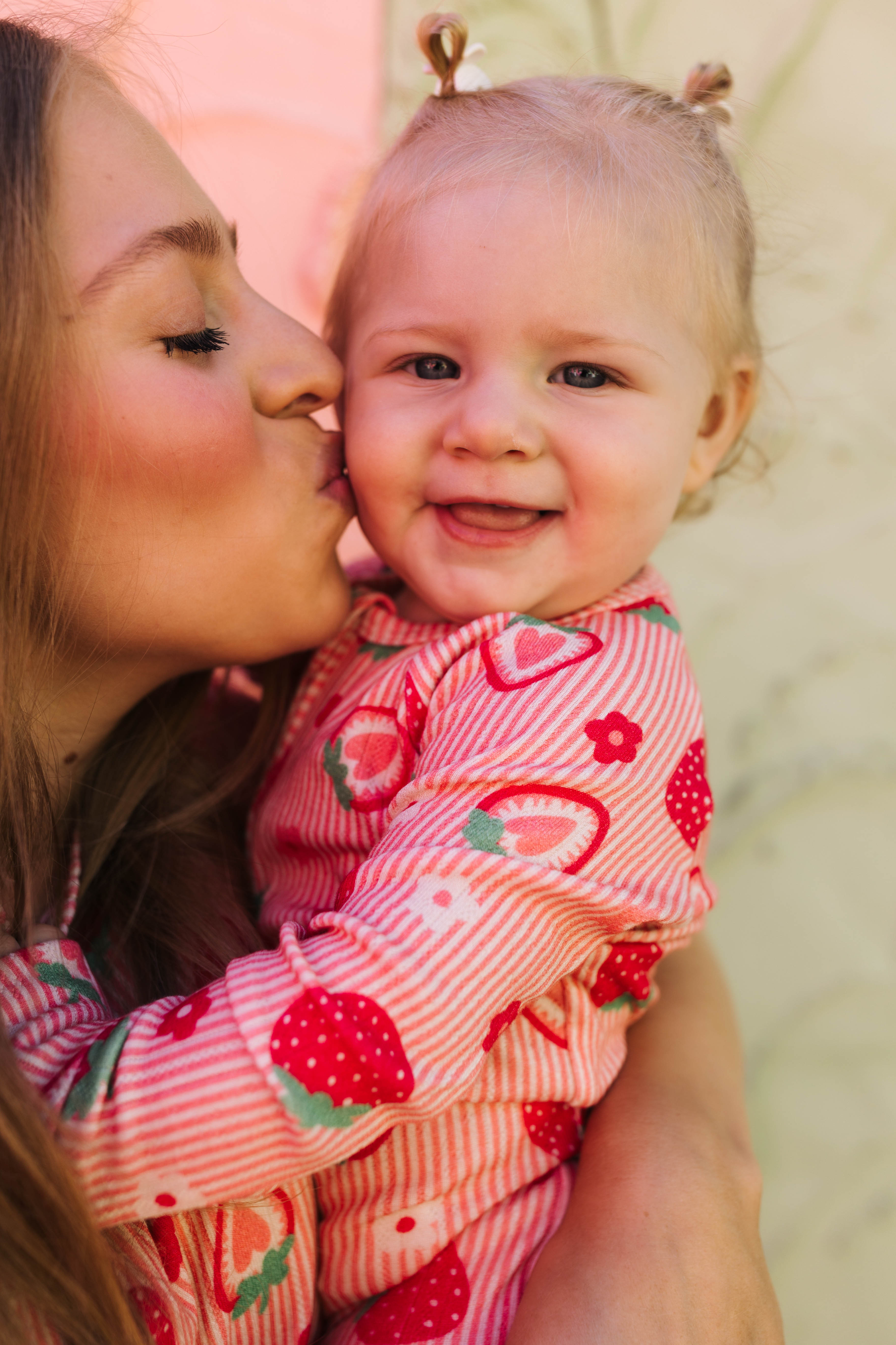 A close-up lifestyle shot of a smiling baby wearing a pink and red strawberry print long-sleeve romper, being kissed on the cheek by a woman.