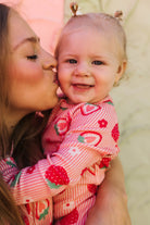 A close-up lifestyle shot of a smiling baby wearing a pink and red strawberry print long-sleeve romper, being kissed on the cheek by a woman.