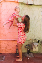 A woman and a baby wearing matching pink heart-patterned pajamas pose together against a textured peach and green stucco wall.