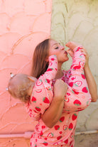 A woman and a baby wearing matching pink strawberry-patterned pajamas pose together against a split pink and beige textured wall.