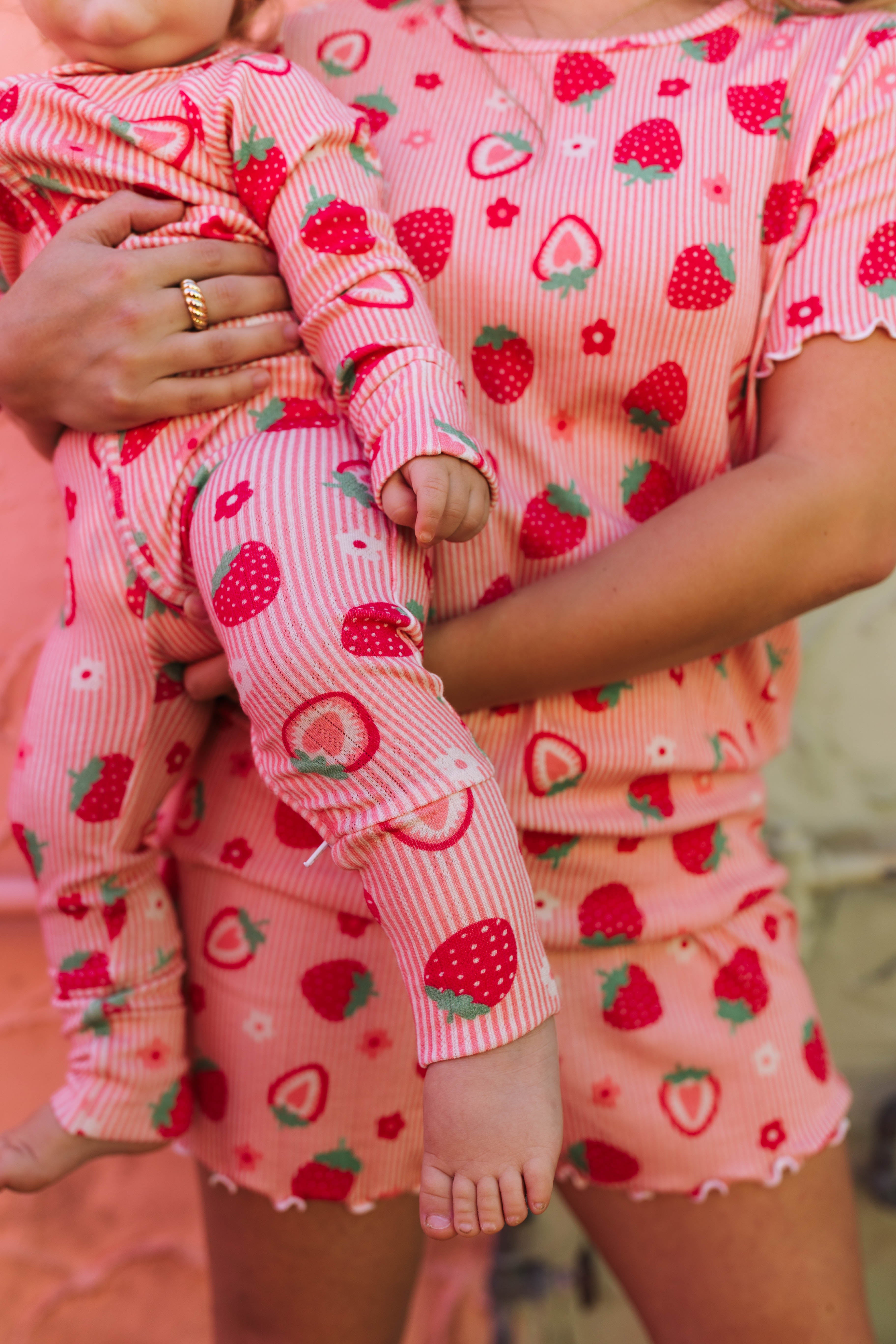 A close-up lifestyle shot of a mother and child wearing matching strawberry print pajamas. The set includes a short-sleeve top and shorts for the adult, and a long-sleeve pajama set for the baby, both featuring a pink and white striped base with red strawberries and green leaves.
