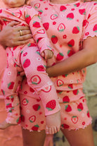 A close-up lifestyle shot of a mother and child wearing matching strawberry print pajamas. The set includes a short-sleeve top and shorts for the adult, and a long-sleeve pajama set for the baby, both featuring a pink and white striped base with red strawberries and green leaves.