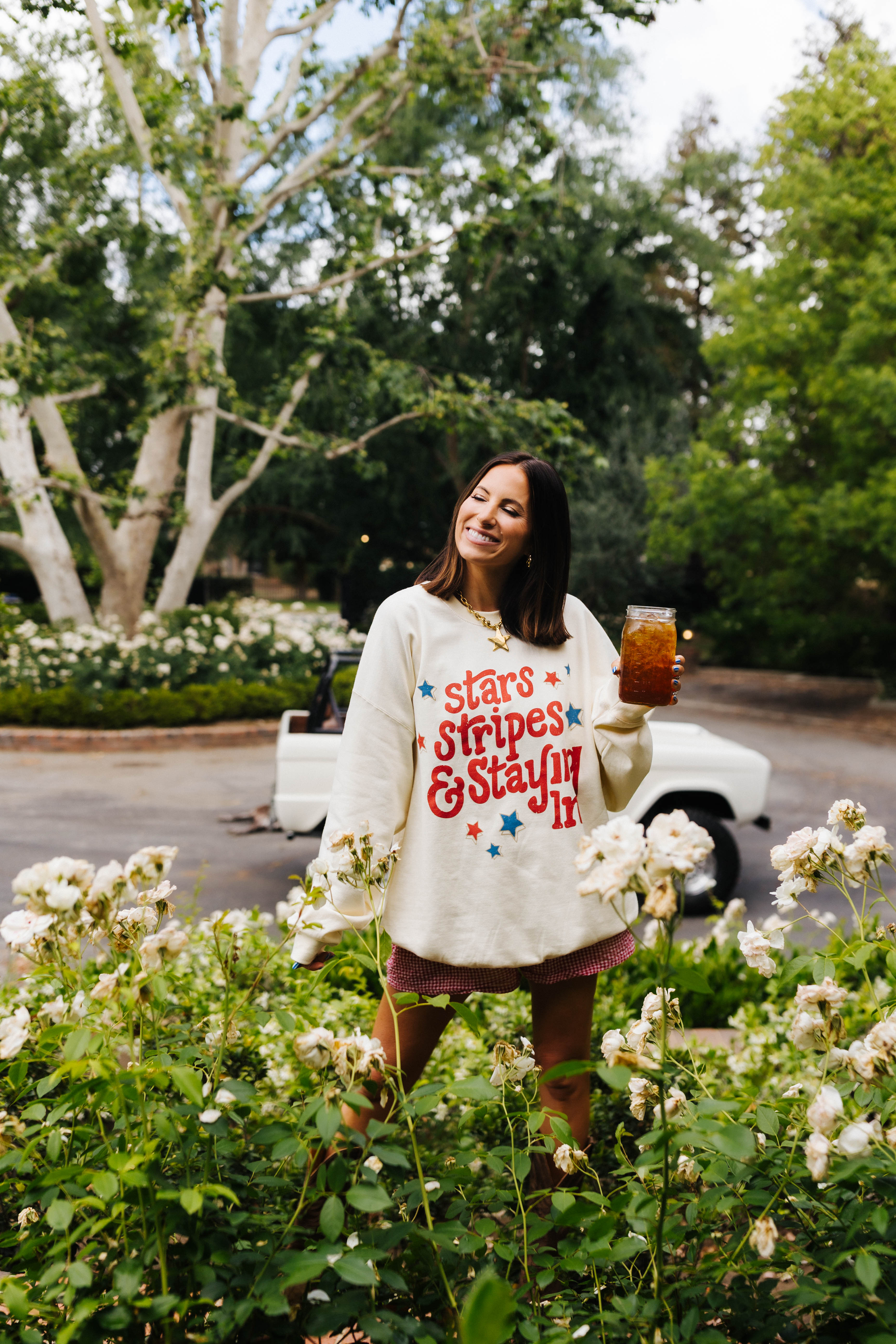Woman in a garden wearing a 'Stars & Stripes & Stayin' It' shirt, holding a drink.