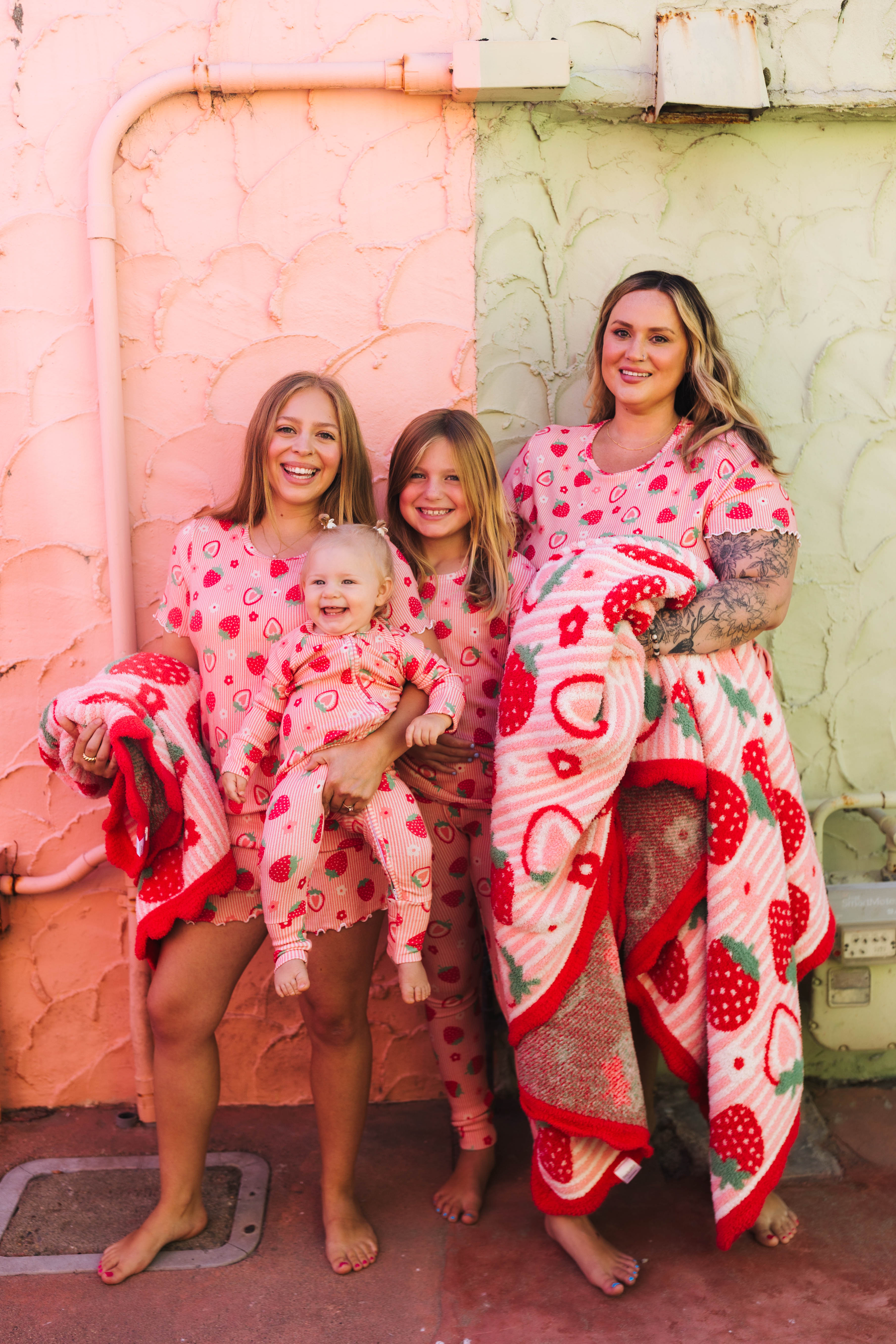 Four girls posing together in matching strawberry-themed pajamas and blankets in front of a split pink and green stucco wall.