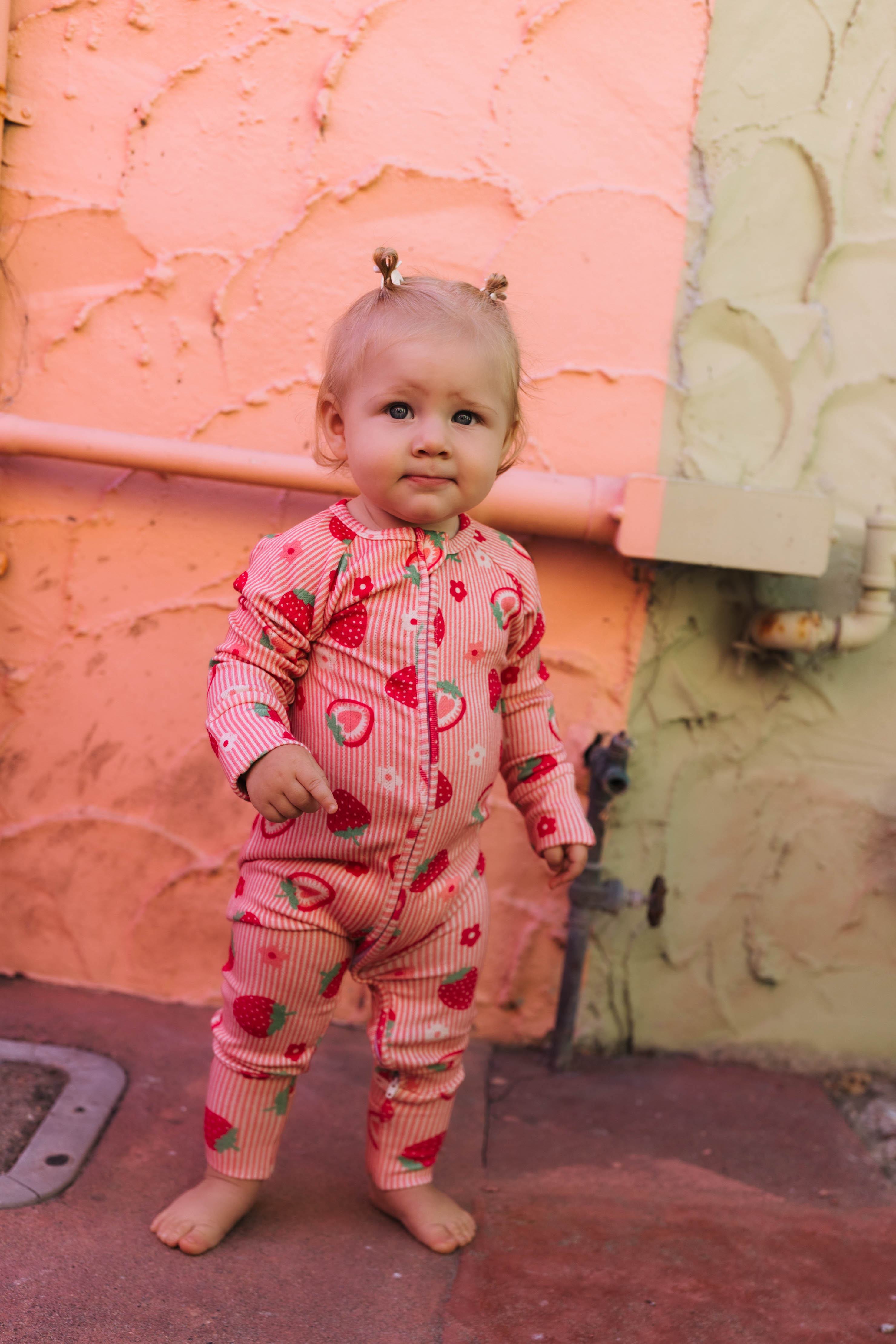 A toddler standing outdoors against a textured peach and beige wall, wearing a pink and red strawberry-and-fruit print one-piece pajama.