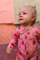 A baby girl wearing a pink and white striped long-sleeve romper with a strawberry and flower print, looking upward outdoors.