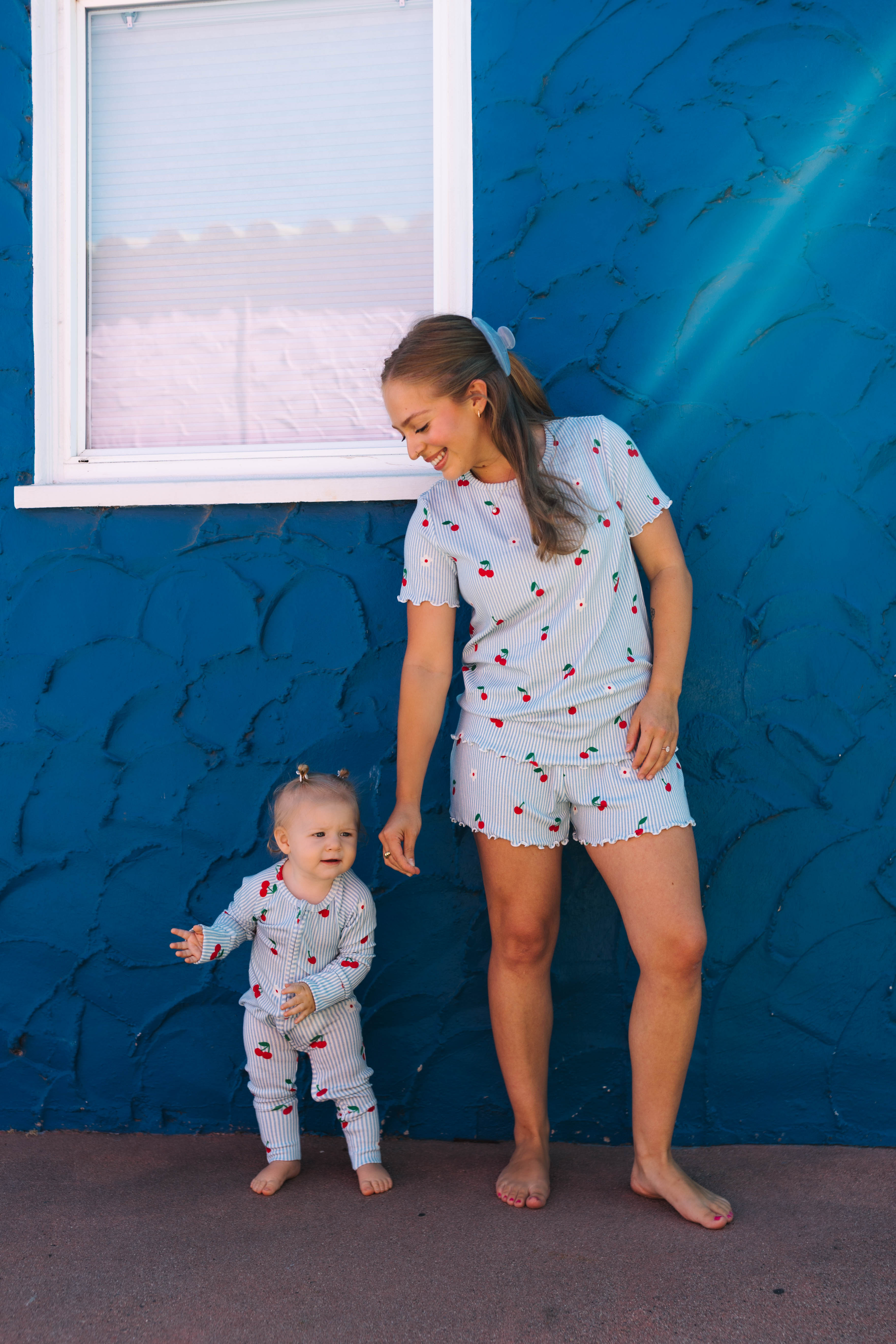 A woman and a toddler wearing matching light blue cherry-print pajamas stand barefoot against a textured blue wall.