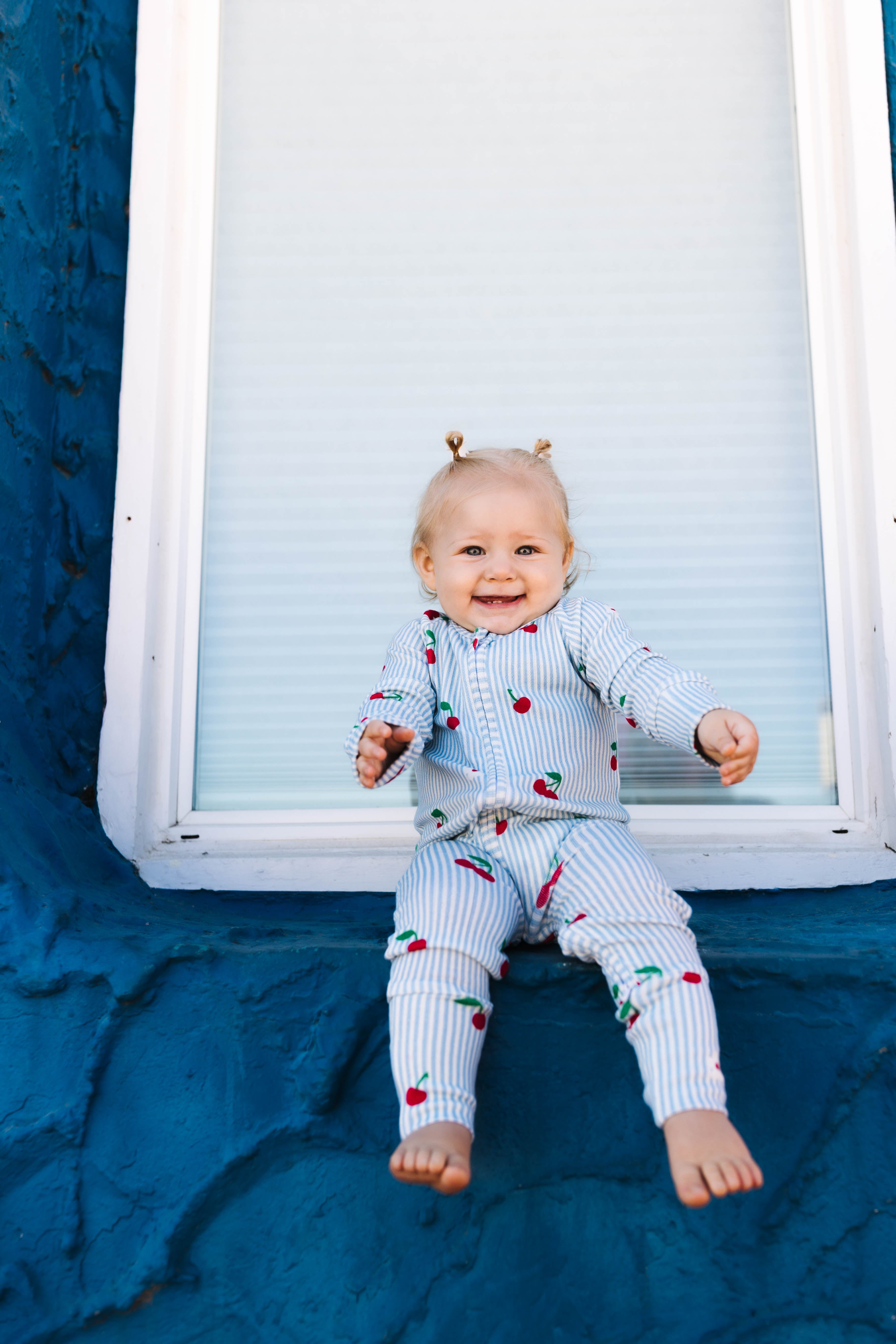 A smiling baby sitting on a dark blue textured surface in front of a white window frame, wearing a light blue and white striped long-sleeve romper and matching pants with a red cherry print.
