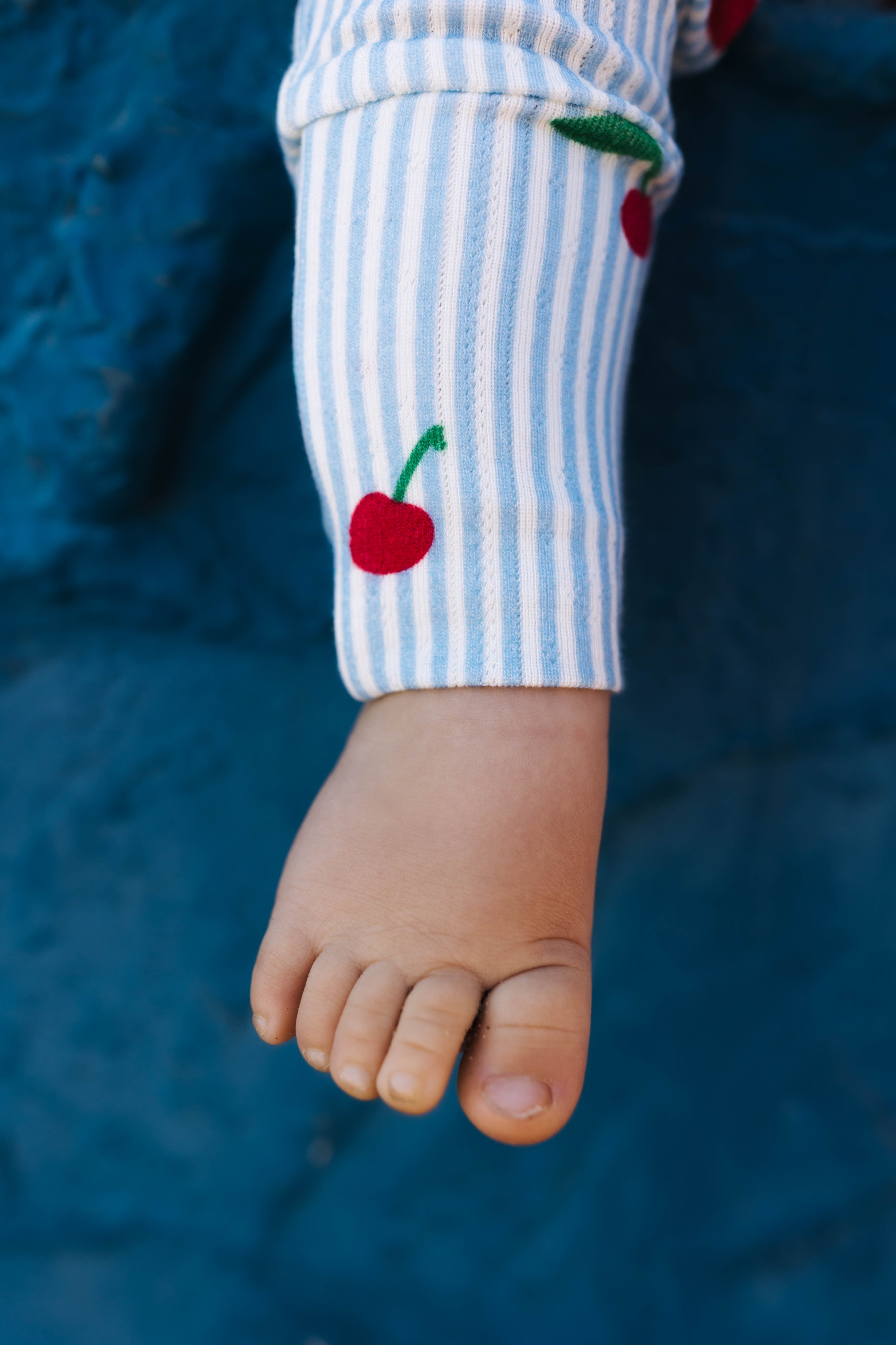 Close-up of a baby's bare foot wearing a light blue and white striped long-sleeve shirt with embroidered red cherries on the sleeve, set against a dark blue textured background.