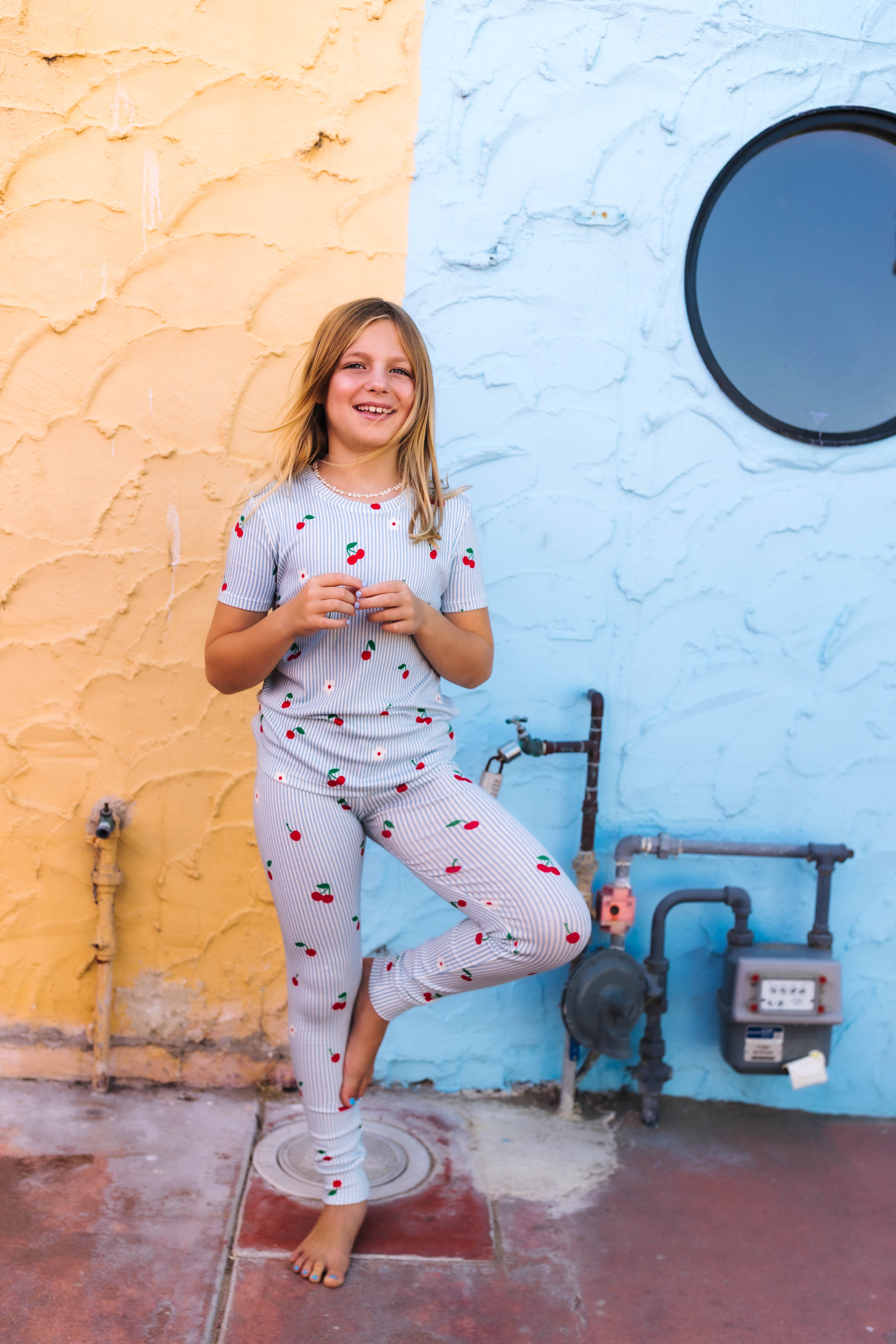 A young girl stands barefoot outdoors against a split yellow and blue wall, wearing a light blue patterned pajama set with small red and white motifs.