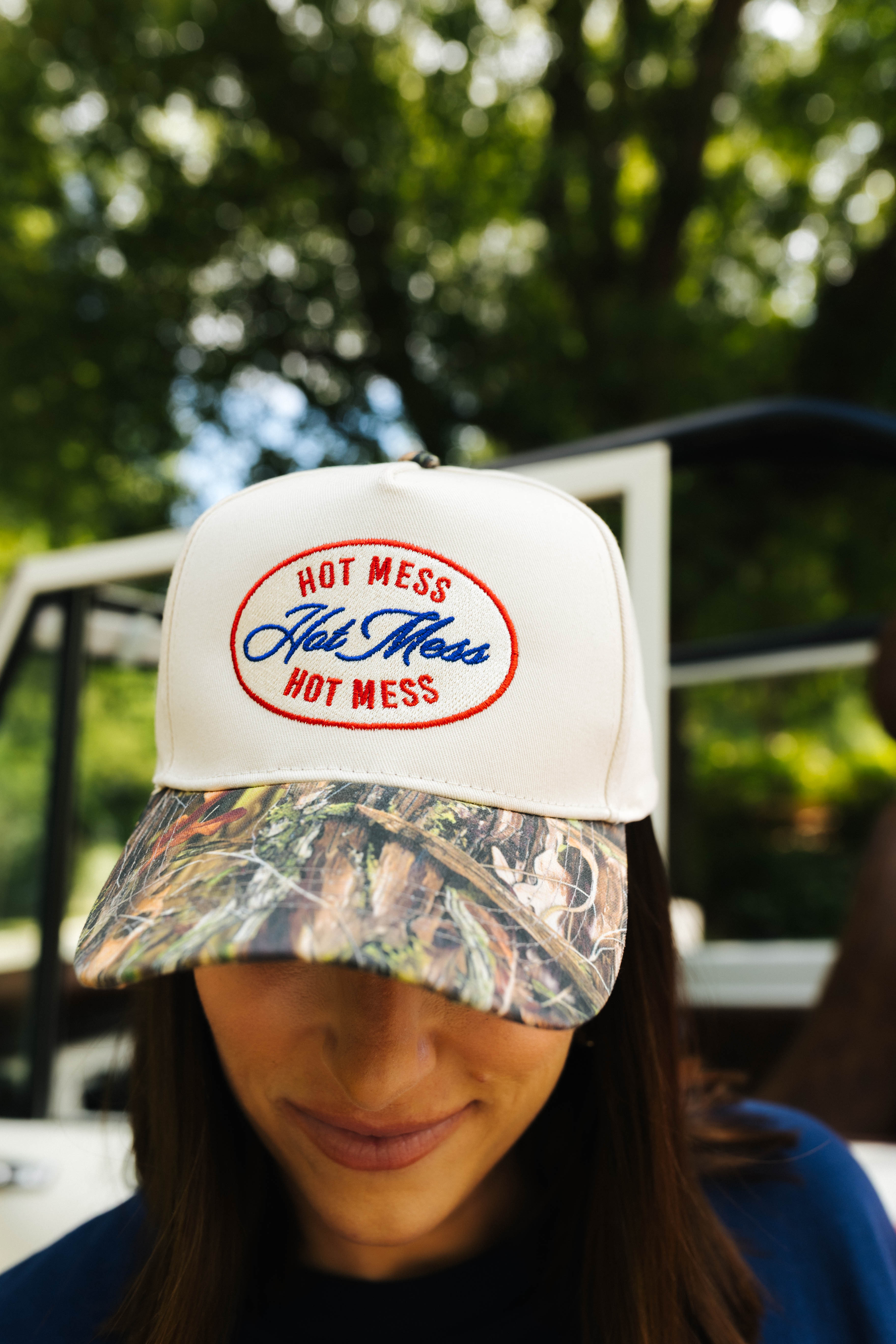 Person wearing a cream cap with a camo bill and 'Hot Mess' branding outdoors