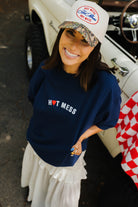 Woman wearing a navy 'Hot Mess' sweatshirt and camouflage cap, standing next to a vintage car.
