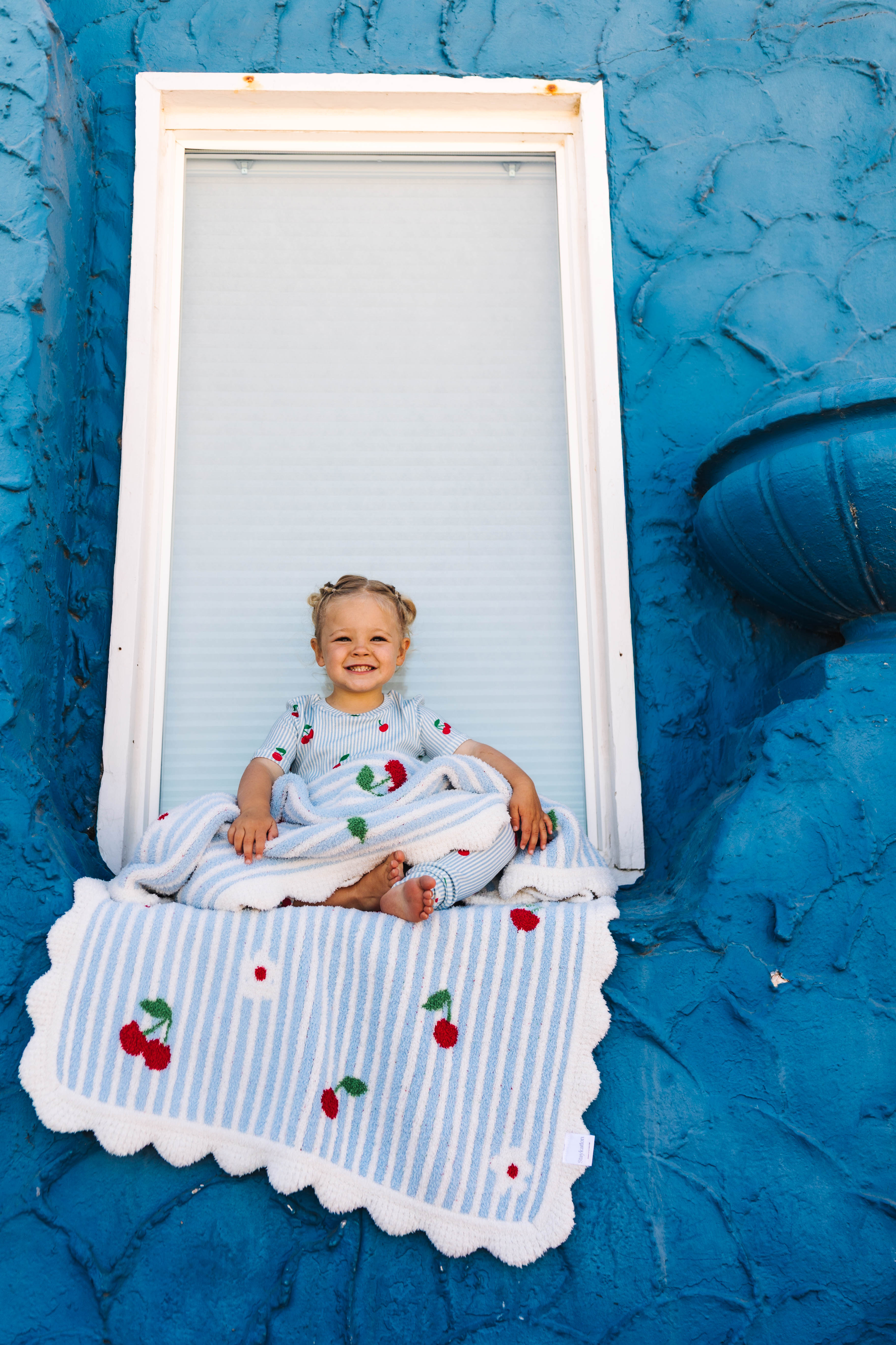 A smiling toddler sits cross-legged on a blue-and-white striped blanket with cherry print, posed in front of a white-framed window on a bright blue textured wall.