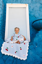 A smiling toddler sits cross-legged on a blue-and-white striped blanket with cherry print, posed in front of a white-framed window on a bright blue textured wall.
