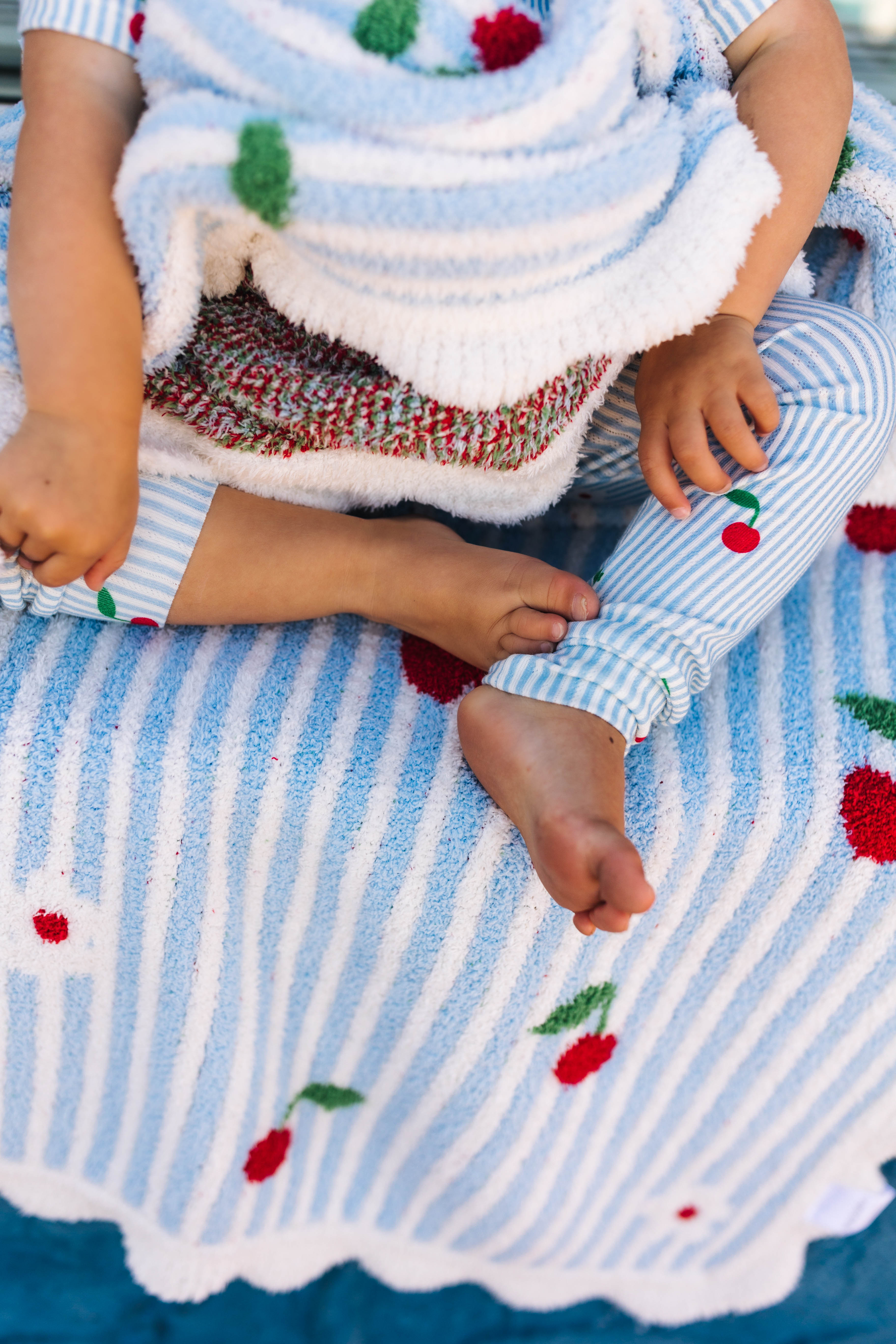 A close-up lifestyle shot of a baby wearing a matching knit outfit consisting of a light blue and white striped pjs with red cherry details and striped pants, sitting on a matching blue and white striped blanket with cherry motifs.