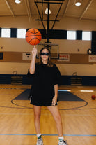 A woman wearing a black oversized t-shirt and black shorts stands on a basketball court, balancing an orange basketball on her finger. She is wearing sunglasses, white socks, and sneakers.